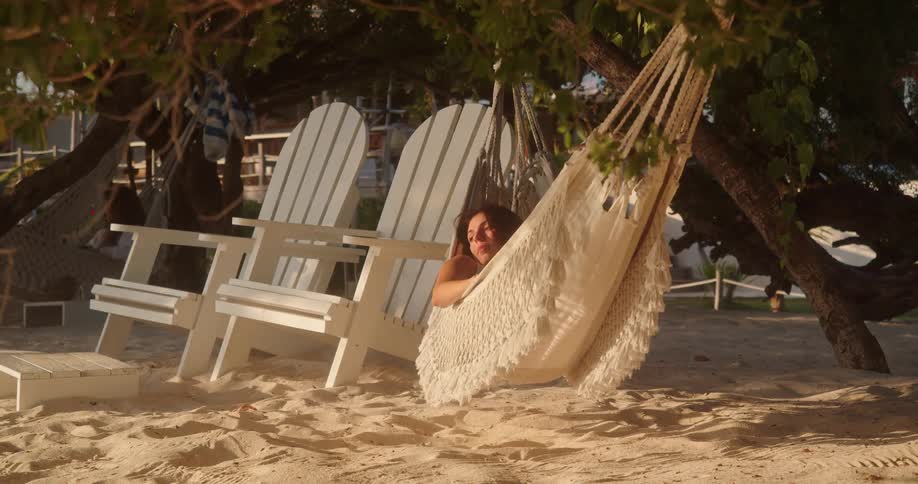 A woman relaxes in a comfortable hammock hanging from a tree on a sandy beach at a tropical resort