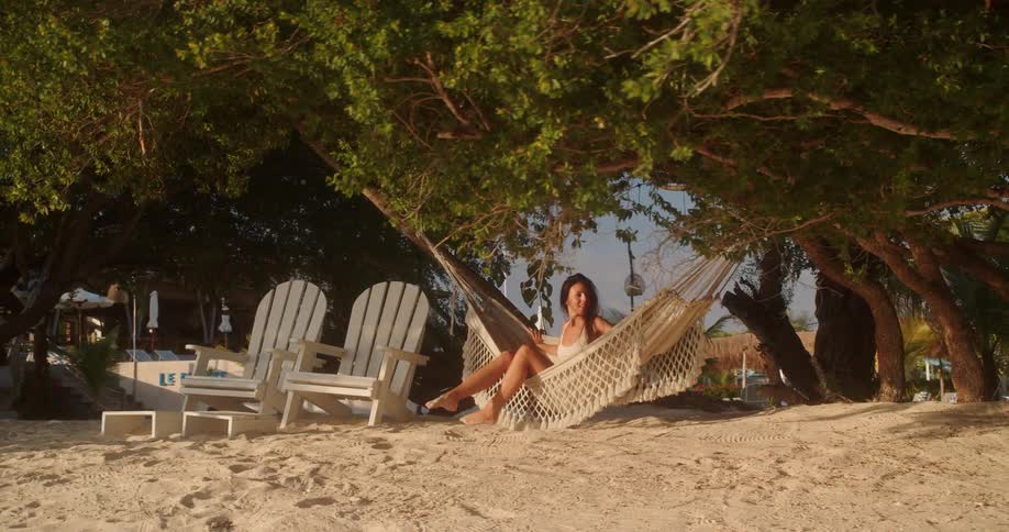 A serene woman in a white bathing suit relaxes in a cozy hammock on a sandy beach, shaded by lush green trees at a tropical resort