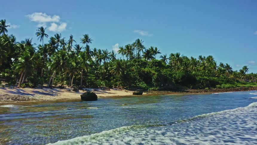 Flying low over the water towards a secluded tropical paradise beach with a sandy shore and palm trees on a sunny day