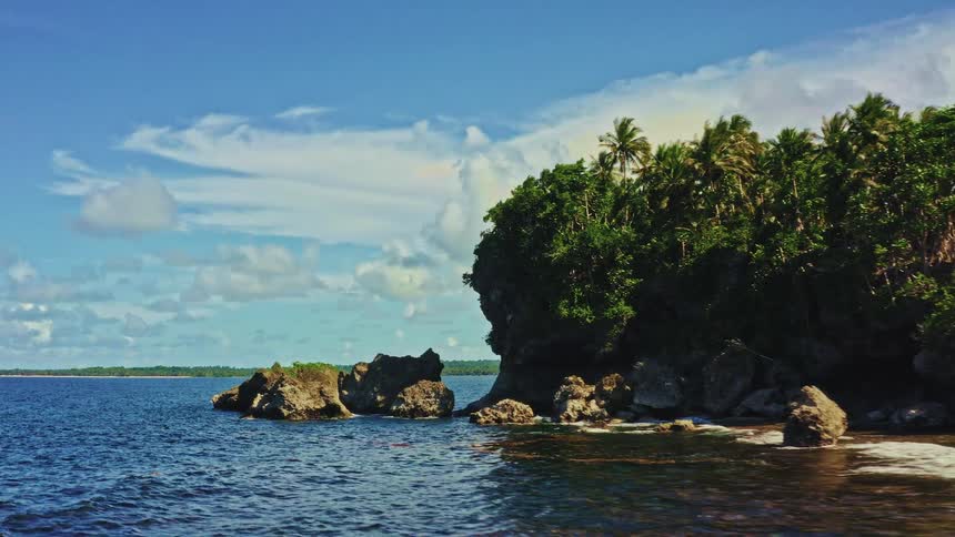 An aerial view slowly glides over the crystal clear ocean towards a rocky, jungle-covered coastline on a beautiful sunny day