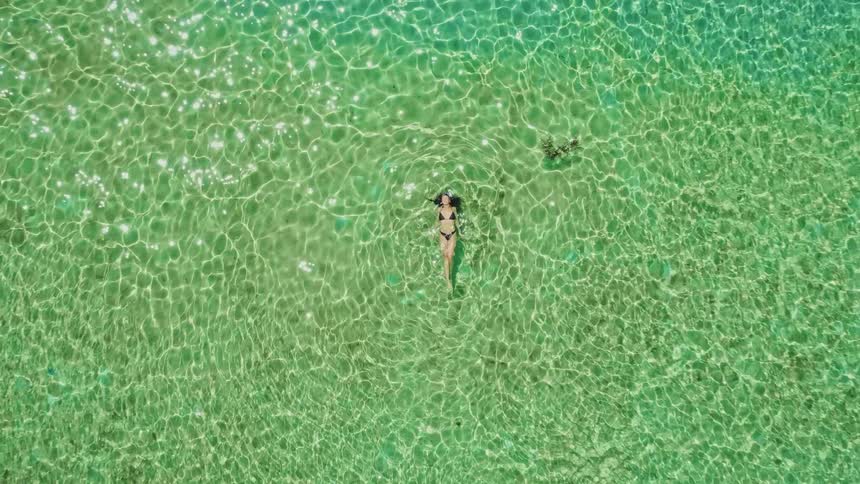 A woman in a black bikini relaxes and floats on her back in the crystal-clear, sun-drenched turquoise ocean water, captured from a top-down aerial perspective that slowly zooms out