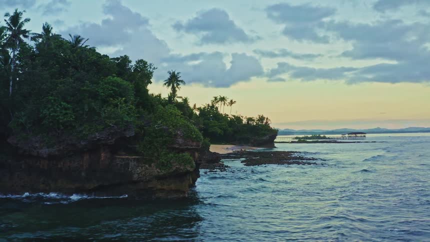 An aerial drone flies along a beautiful, rocky tropical shoreline revealing a hidden sandy beach at sunrise