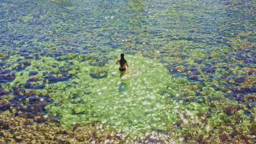 A drone follows a woman in a bikini as she wades through the stunning, sun-drenched shallow waters of a tropical sea