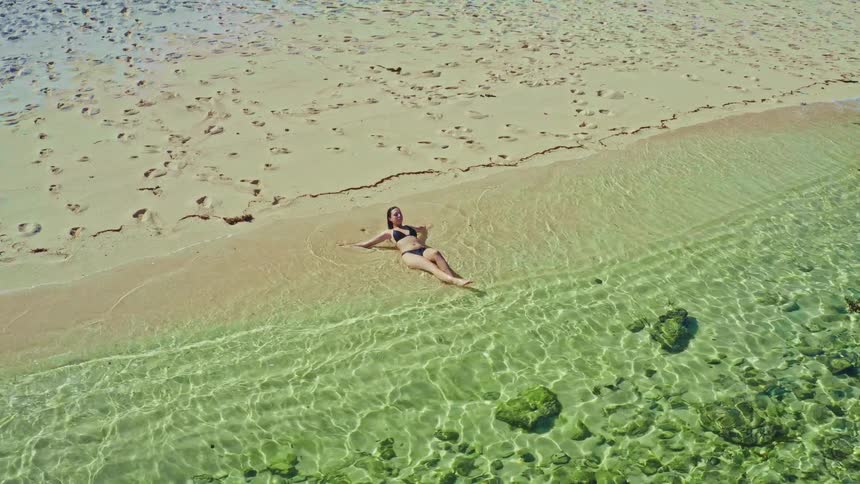 An aerial view of a woman in a bikini relaxing on a sandy beach by the clear turquoise ocean