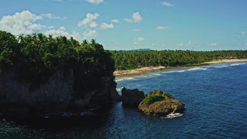 An idyllic aerial view slowly approaches a jungle-covered rocky cliff next to a beautiful tropical beach and deep blue ocean
