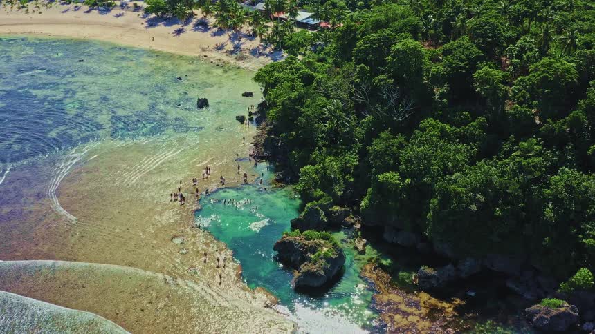 An aerial view captures the vibrant turquoise waters of a natural rock pool where people swim along a lush tropical coastline
