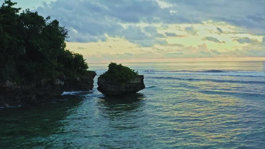 A slow aerial pan across a tropical, rocky coastline revealing the calm ocean waters at sunset with ships on the horizon