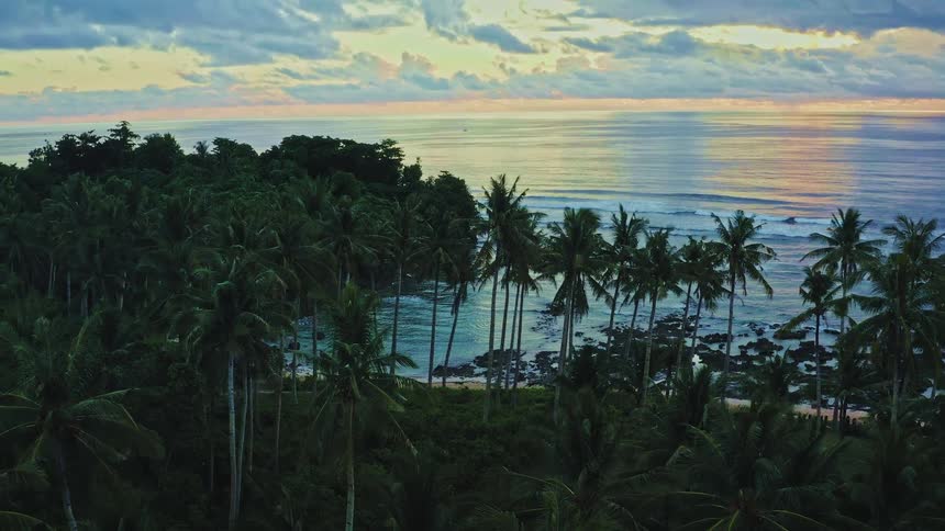 An expansive aerial shot reveals a stunning tropical island cove lined with palm trees during a colorful sunset