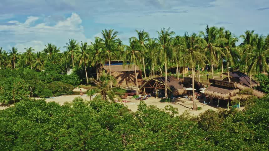 Aerial view reveals a secluded tropical beach resort with thatched-roof huts nestled among palm trees, where people are relaxing and swimming in a calm lagoon