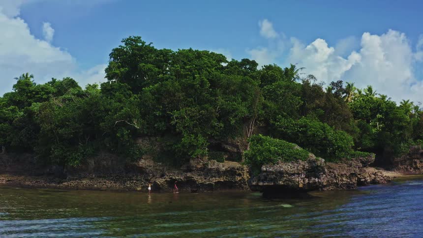 Swimmers enjoy the tranquil, shallow waters beside a rocky, jungle-covered tropical coastline on a bright sunny day
