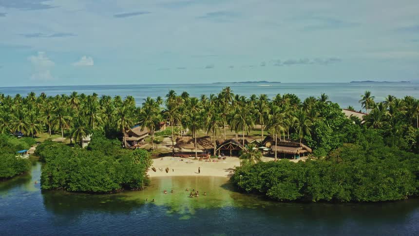 An aerial shot pulls back from a woman waving on a sandy beach to reveal a beautiful tropical resort island with palm trees, thatched huts, and clear blue water