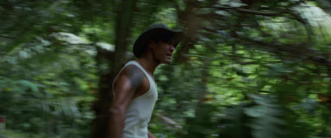 A man in a hat runs with a determined look on his face through a dense green jungle