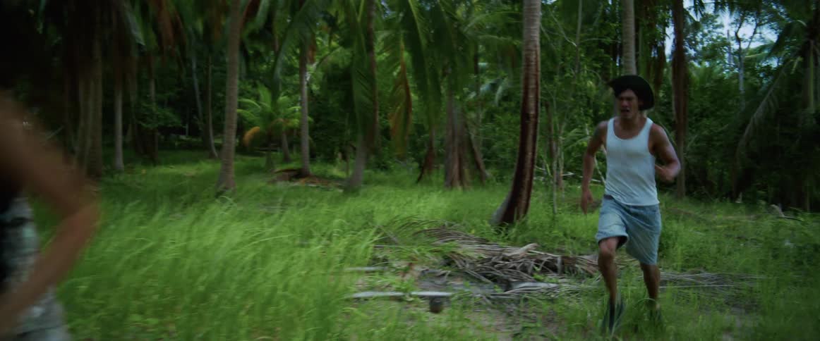 A frightened woman runs away from a man chasing her through a dense tropical jungle in slow motion