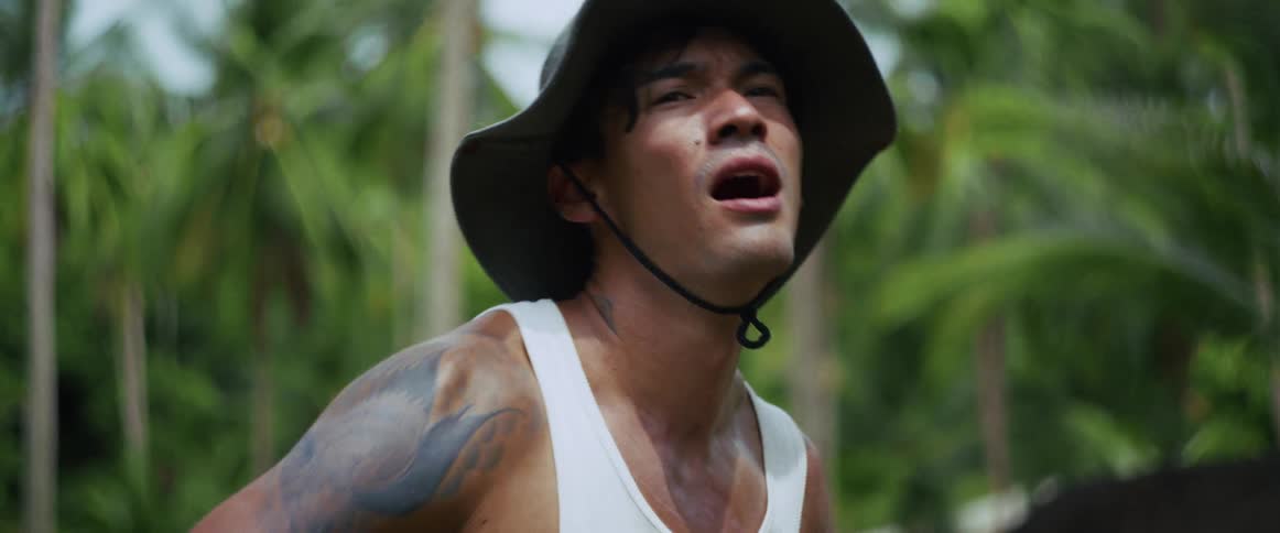 A sweaty, muscular man in a hat breathes heavily and looks up, exhausted from hard work in a lush tropical jungle