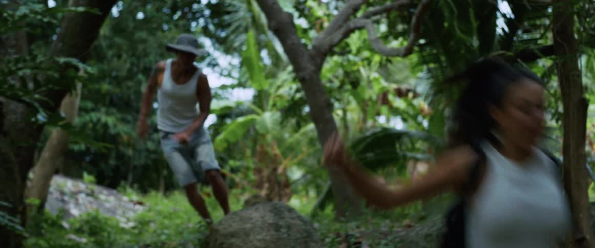 A man and woman run in slow motion through a dense, green tropical jungle