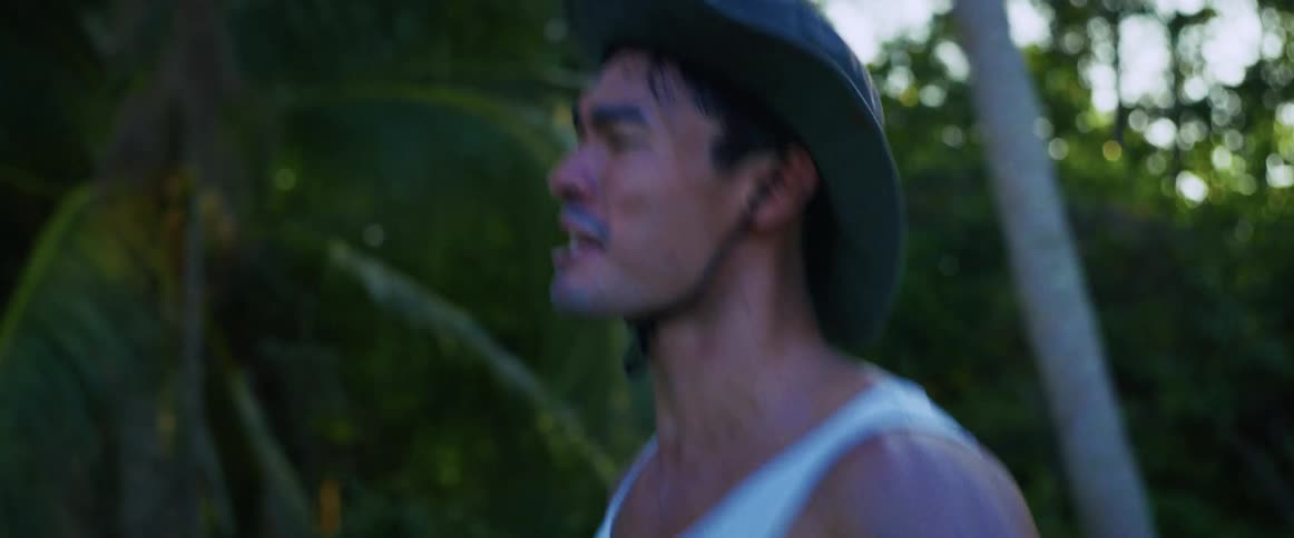 An intense close-up shot of a sweaty man in a hat and tank top running desperately through the jungle