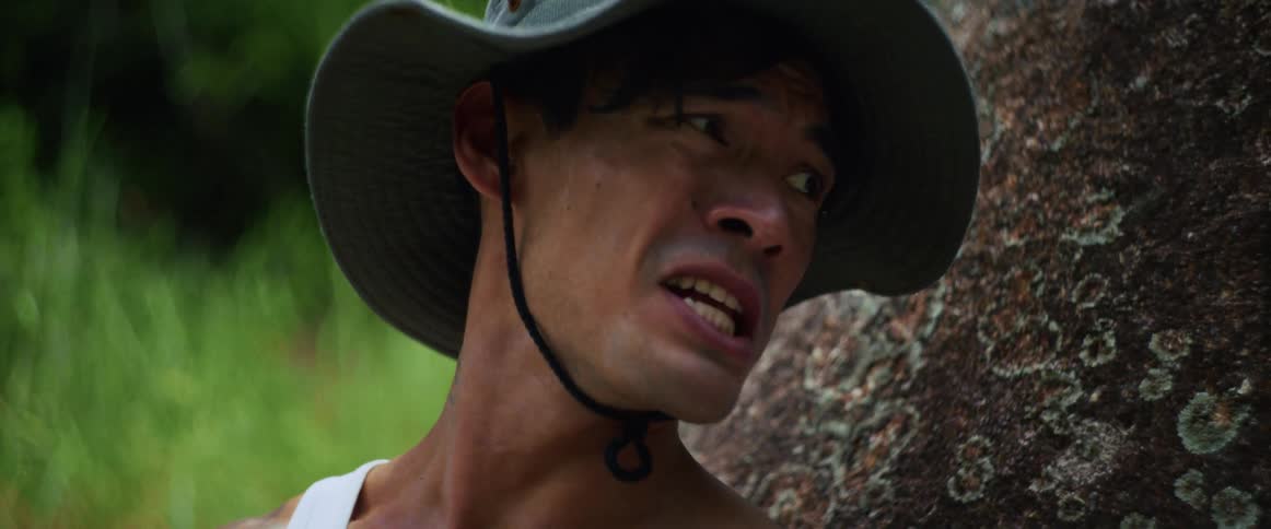 A close-up of a man in distress, breathing heavily and looking fearful while leaning against a rock in the jungle