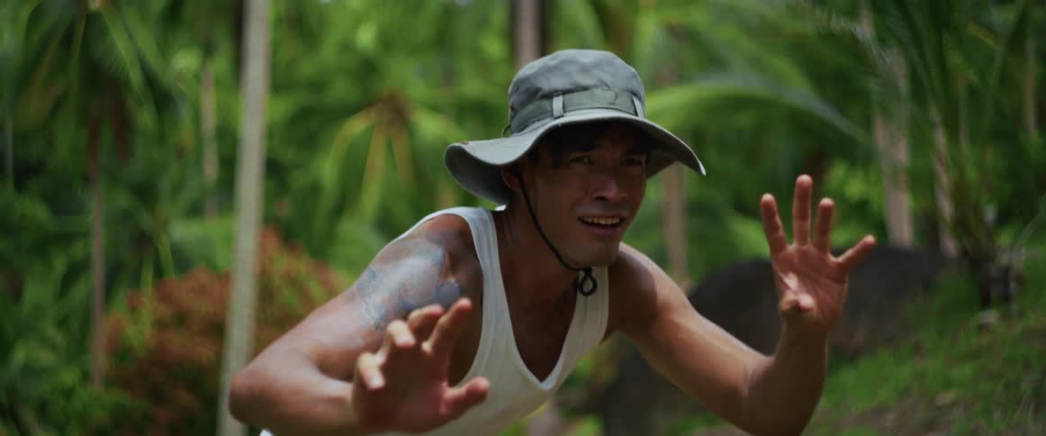 A man with a tattoo wearing a bucket hat looks scared and holds his hands up defensively in a lush green jungle