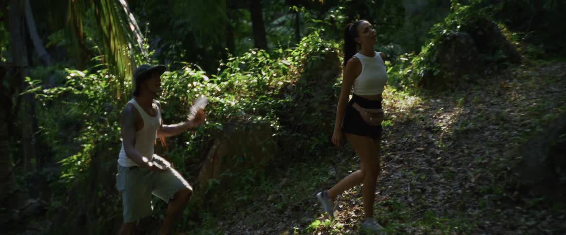A man cools a woman with a hand fan while they hike uphill through a lush, sun-dappled tropical jungle