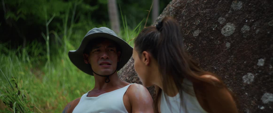 An exhausted couple takes a break from hiking, resting against a rock in the dense, humid jungle