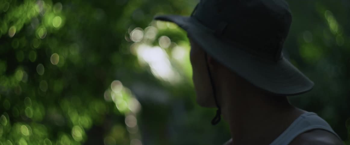 Close-up of a sweaty and exhausted man in a hat looking lost and disoriented in a dense jungle in slow motion
