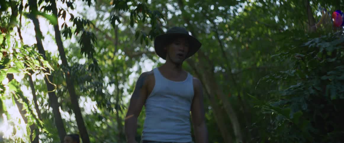 A couple cautiously explores a dense, sun-dappled jungle, stopping to look around with wary and surprised expressions
