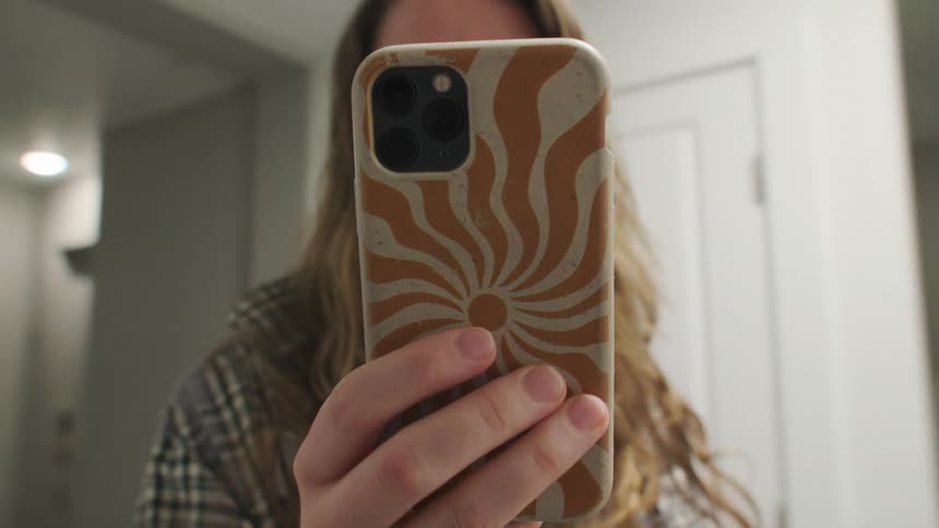 A mysterious person films themselves in a mirror while walking backward down a hallway, their face obscured by their phone
