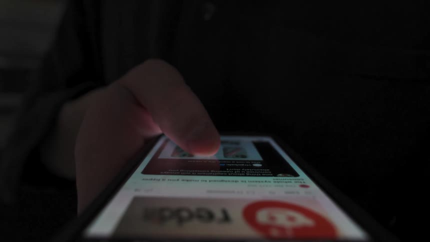 A close-up, low-angle shot of a person's thumb scrolling through a social media feed on a smartphone at night