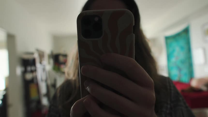 A person holds a phone up to a mirror, filming their reflection as they walk backward through the rooms of their home