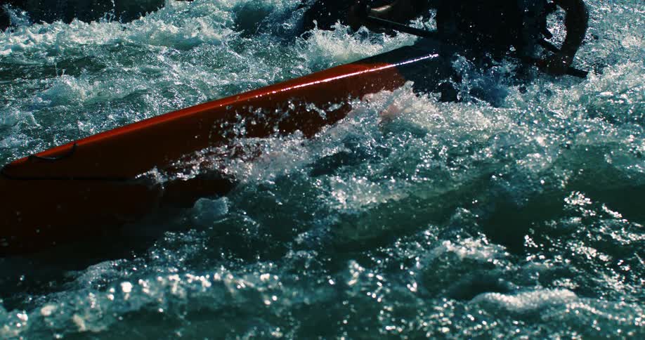 A close-up view shows an orange kayak navigating through turbulent whitewater rapids in dramatic slow motion