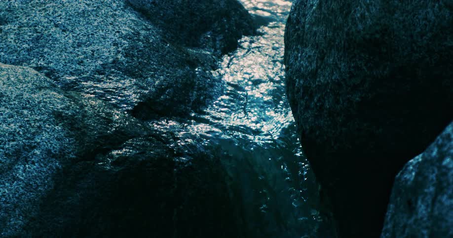 A dark and moody close-up of a clear stream of water flowing between textured rocks in super slow motion