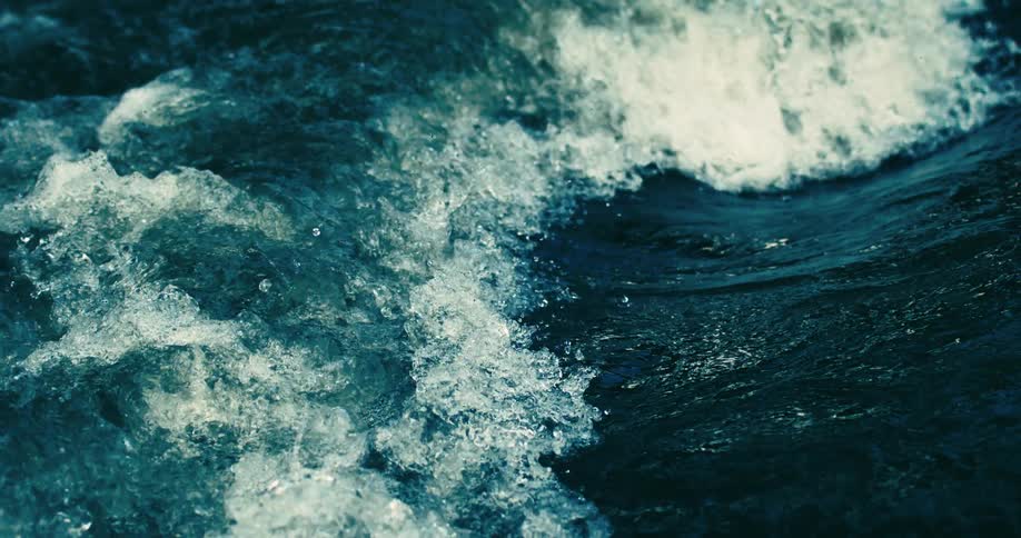 A dramatic slow-motion close-up captures the powerful churning of dark blue water as it creates white foamy rapids