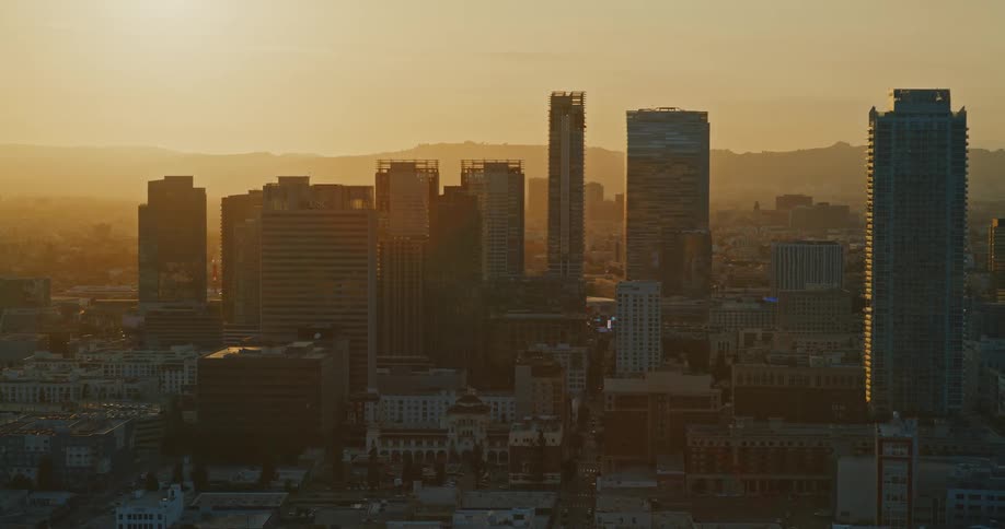 An aerial view of the hazy downtown Los Angeles skyline with skyscrapers silhouetted against the setting sun during golden hour