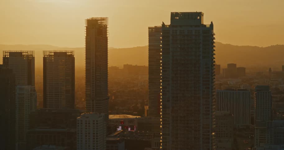 A slow push-in aerial shot captures the silhouetted skyscrapers of a downtown city glowing against a hazy, golden sunset