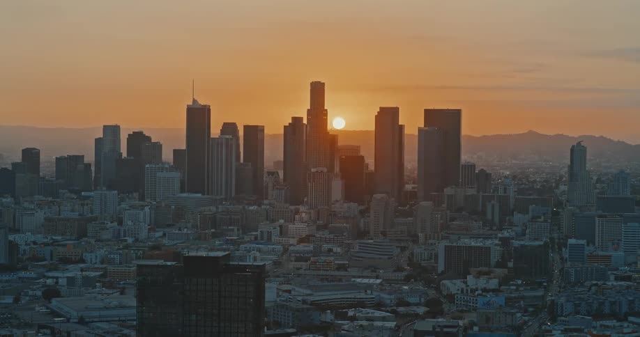 A serene aerial view shows the sun setting behind the downtown Los Angeles skyline, bathing the cityscape in a warm, golden light