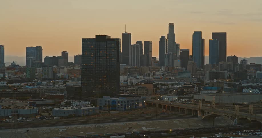 An aerial view slowly reveals the downtown Los Angeles skyline, iconic bridges, and industrial areas bathed in the warm glow of a beautiful sunset