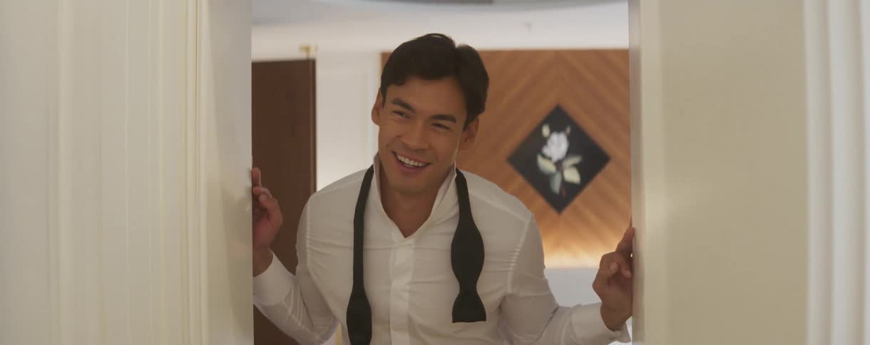 A charming man in a white dress shirt and untied bow tie smiles lovingly as he stands in a doorway