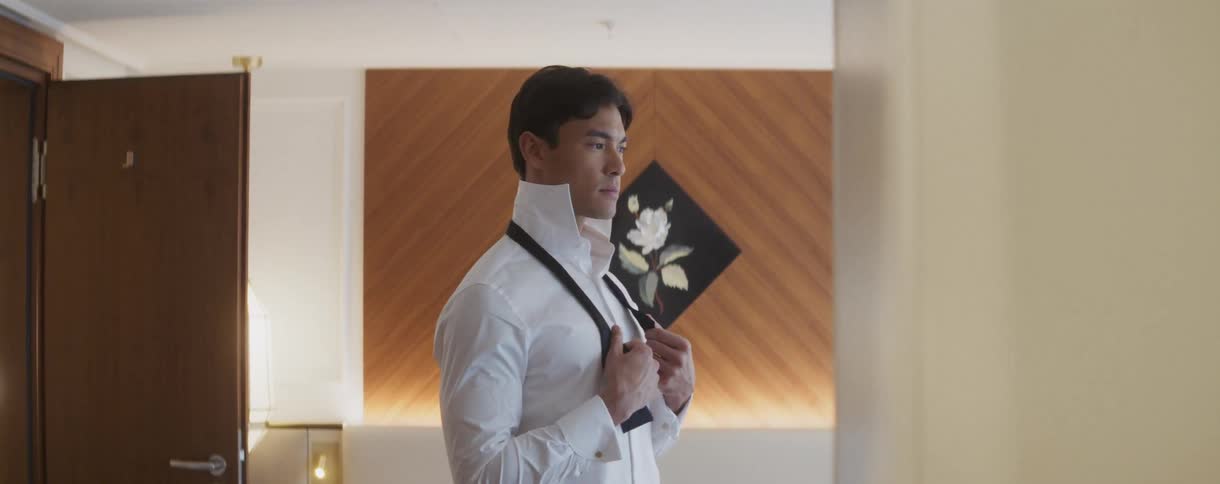 A sophisticated man stands in a stylishly lit room, thoughtfully preparing to tie his bow tie for a formal event