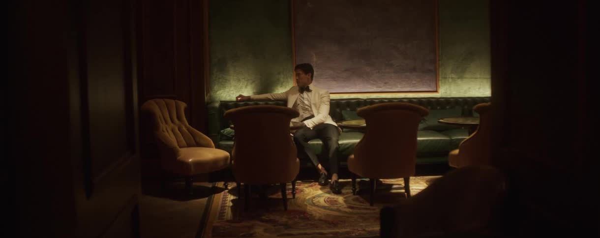 A stylish young man in a white dinner jacket sits alone and waits in a moody, upscale lounge