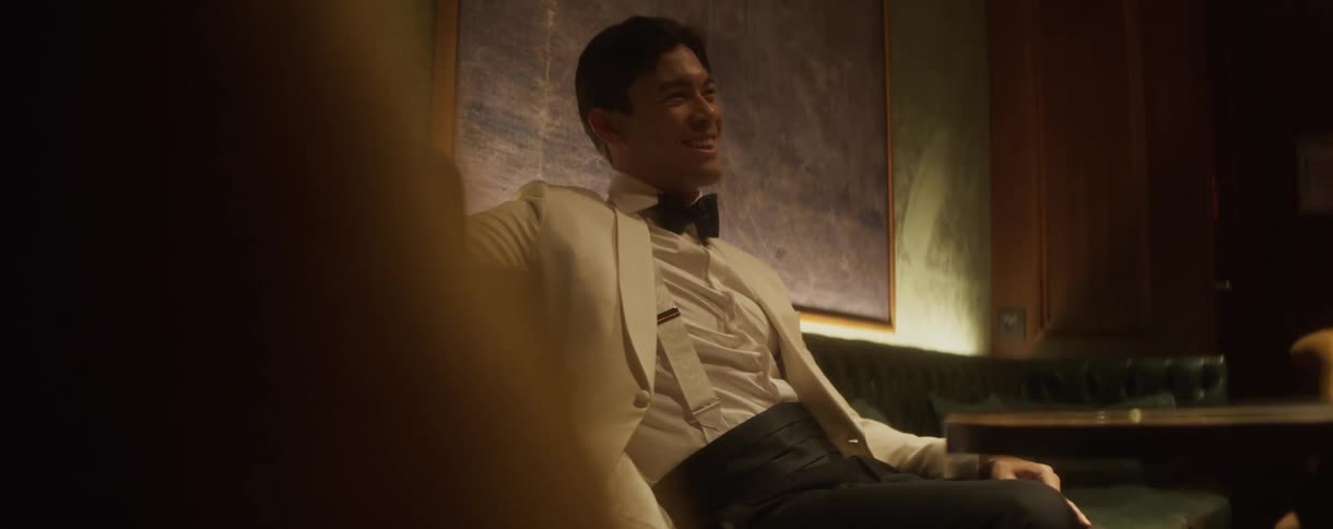A suave and handsome man in a white tuxedo jacket and bow tie smiles while seated in an exclusive, dimly lit lounge