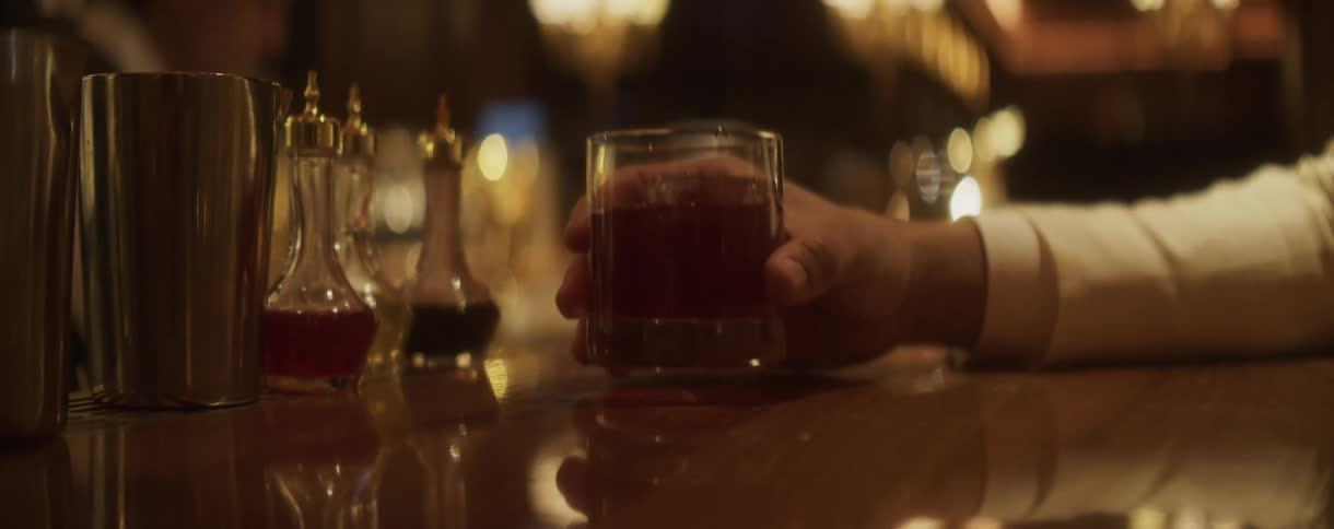 A man's hand slowly picks up a red cocktail with an ice cube from a polished wooden bar in a dimly lit, elegant lounge