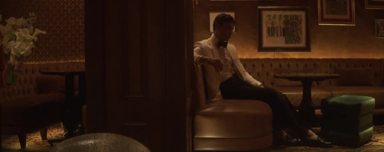 A well-dressed man sits alone in a luxurious, dimly lit lounge, looking pensive and mysterious
