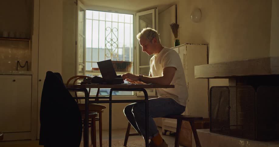 A man with white hair sits at a table in a sunlit room and begins working on his laptop