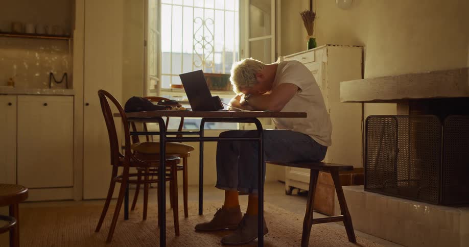 A tired freelancer working on a laptop at a sunlit dining table lays his head down in exhaustion