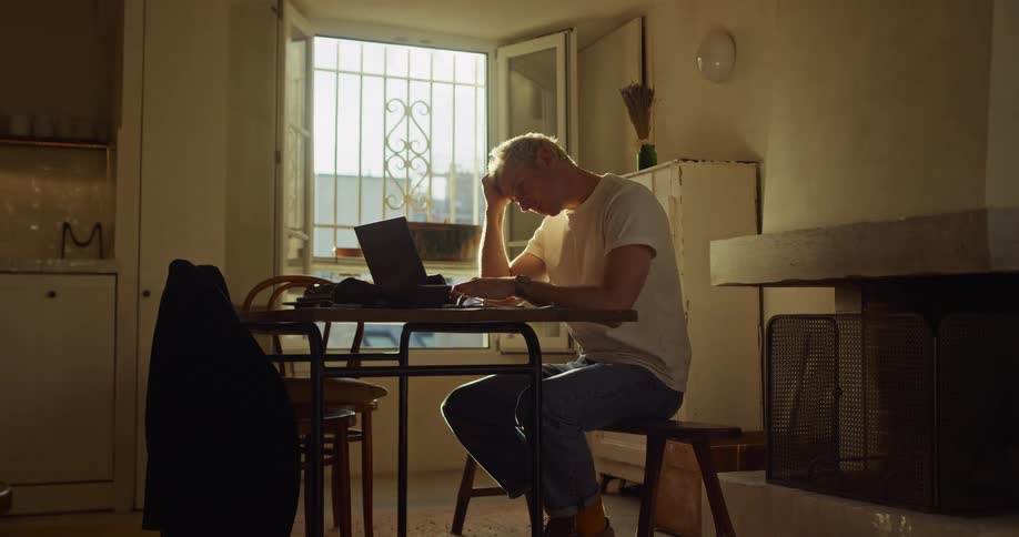 A stressed man with bleached blonde hair works on his laptop in his apartment, illuminated by warm sunlight from a window