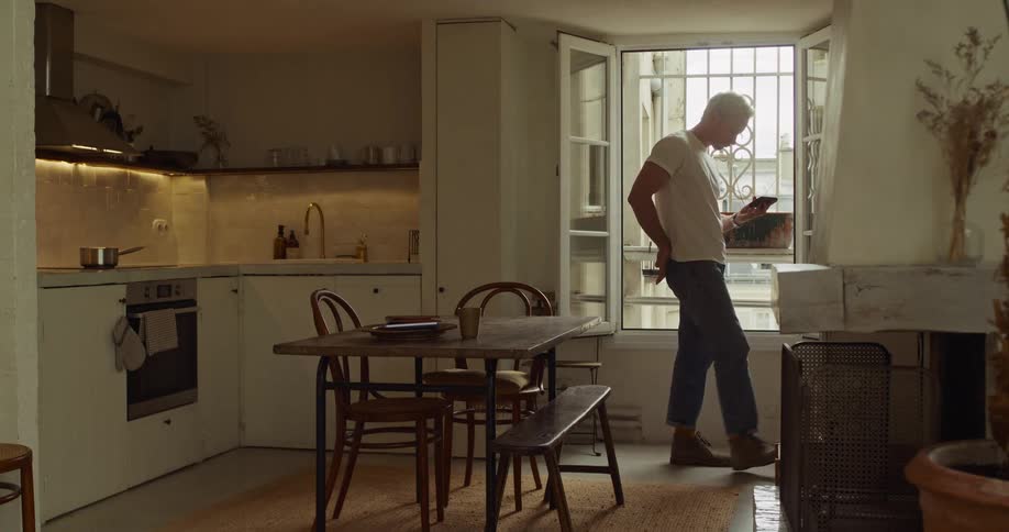 A man casually uses his smartphone while standing by the window in his rustic, light-filled apartment