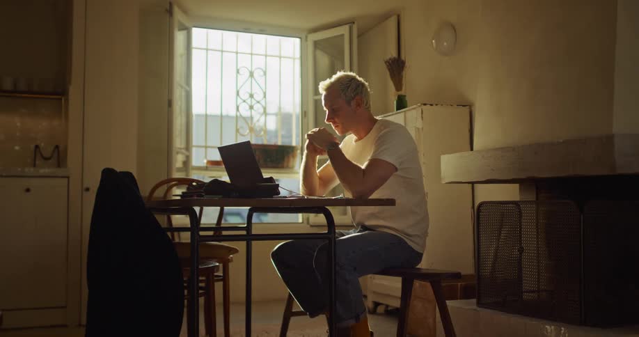 A serious young man with bleached blonde hair contemplates his work on a laptop in a sunlit, rustic apartment