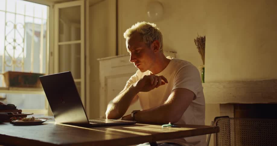 A focused young man with bleached blonde hair sits at a table and participates in a video call on his laptop in a sun-drenched, rustic room