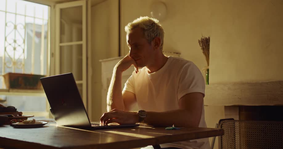 A focused man with blond hair works from home on his laptop, sitting at a wooden table in a room bathed in warm, golden hour sunlight