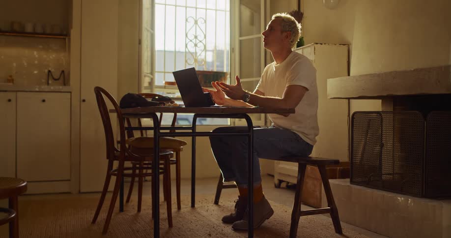 A pensive young man with blonde hair works on his laptop at a rustic table, pausing to think in a sunlit room